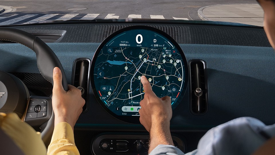 MINI Électromobilité – écran OLED central avec navigation. Conducteur et passager interagissant avec l’écran d’infodivertissement circulaire MINI affichant la navigation.